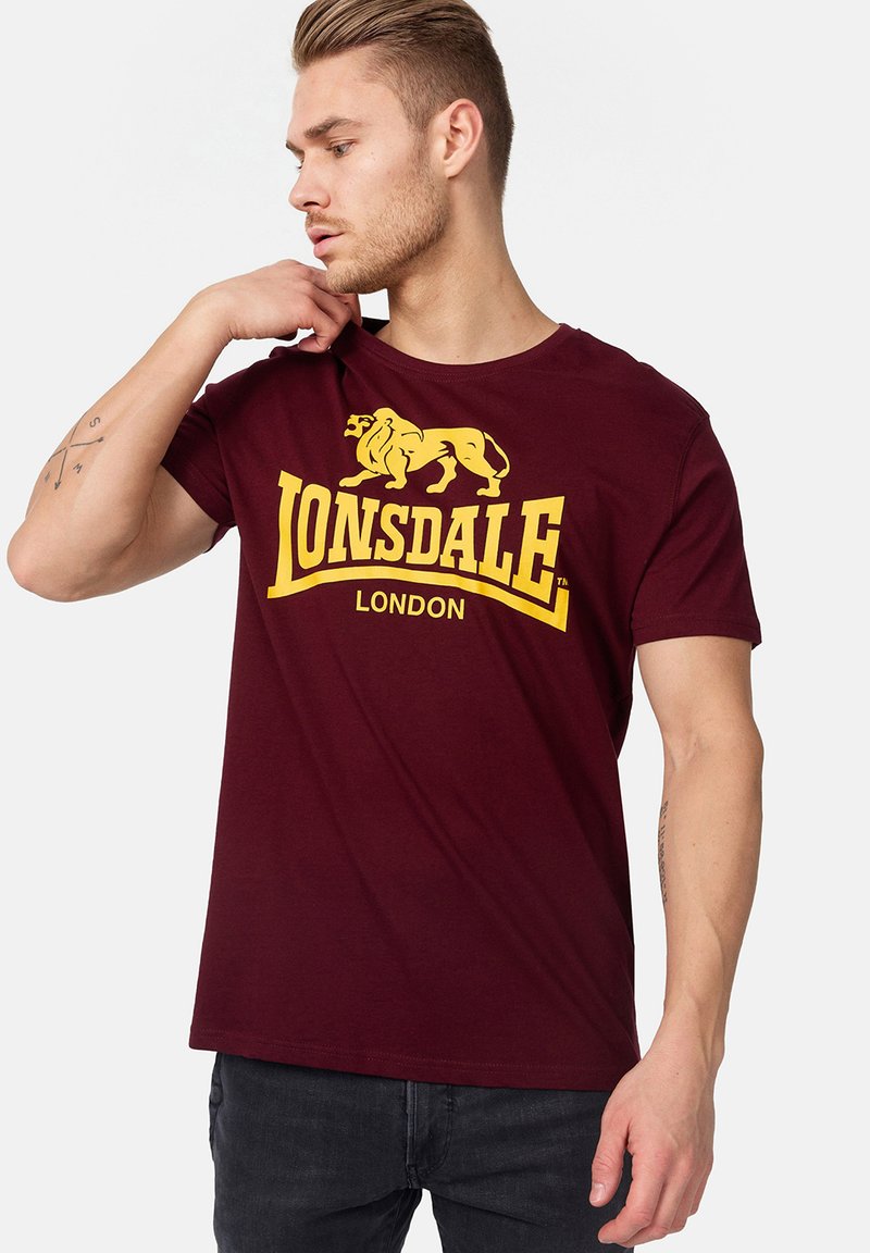 Burgundi színű póló sárga oroszlán logóval és "LONSDALE LONDON" felirattal. Rövid ujjú és kerek nyakú. Pamut anyag, laza fazon.