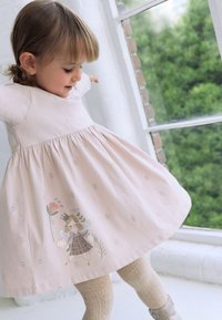 Ružové šaty s krátkym rukávom a rozšírenou sukňou, zdobené vyšívanými kvetinovými detailmi a postavičkou na leme. Pletené krémové pančuchy.