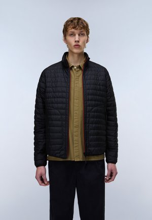 Jong persoon draagt een zwart doorgestikt jack over een olijfgroen overhemd met knopen en zwarte broek, staand tegen een effen lichte achtergrond.