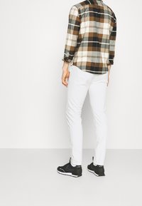 Camicia in flanella a quadri nei toni del marrone, crema e nero; pantaloni bianchi slim-fit; scarpe da ginnastica nere. Maniche arrotolate, texture a motivo visibile.