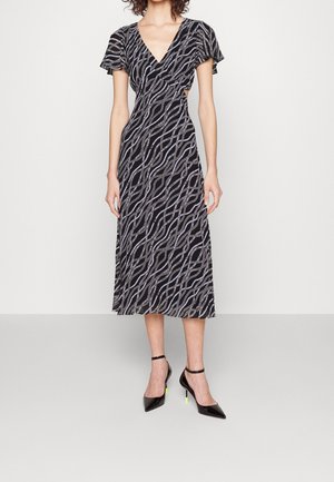 Woman wearing a black midi dress with white swirling chain patterns, short flutter sleeves, v-neck, and black pointed heels with ankle straps.