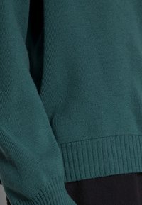 Türkiser gestrickter Pullover mit geripptem Saum und Bündchen, der über eine weiche Textur und einen lockeren Schnitt verfügt. Fokus auf die Details der Ärmel und Seiten.