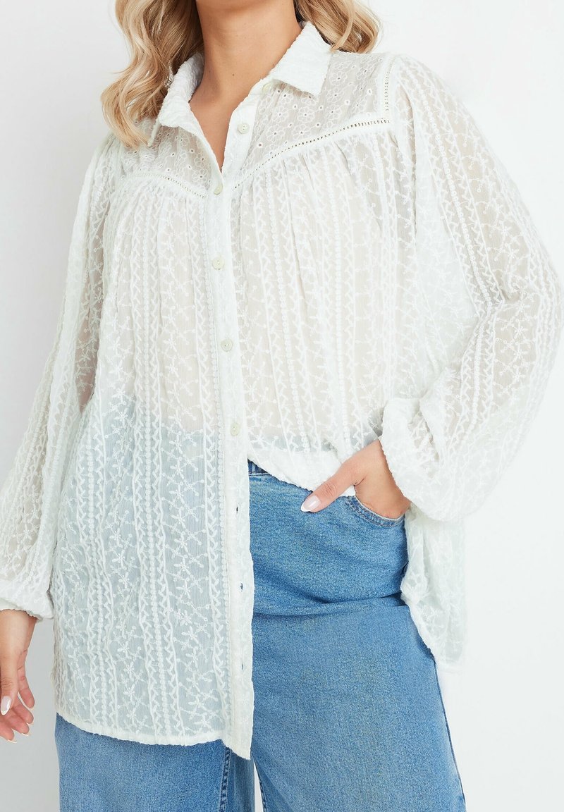 Femme portant un chemisier blanc transparent brodé à manches longues avec des boutons, associé à un jean en denim bleu, main dans la poche.