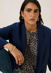 Blazer marine à coupe décontractée, porté par-dessus un blouse à motif léopard. Les accessoires incluent plusieurs bracelets et des boucles d'oreilles créoles.