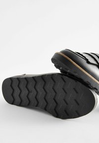 Schwarze Fußbekleidung mit einer strukturierten Gummisohle, die ein Zickzackmuster aufweist, gestaltet mit geschichteten Trägern über dem Obermaterial.