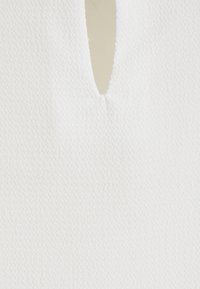 Tissu blanc texturé avec une découpe en forme de goutte d'eau au niveau de l'encolure. La surface présente un motif subtil, ajoutant un intérêt visuel sans brillance.