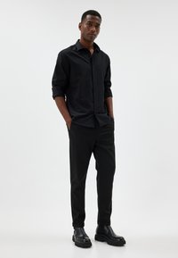 Camicia nera con bottoni e maniche arrotolate, abbinata a pantaloni neri e stivaletti neri. Tessuto liscio, vestibilità su misura, design minimalista.