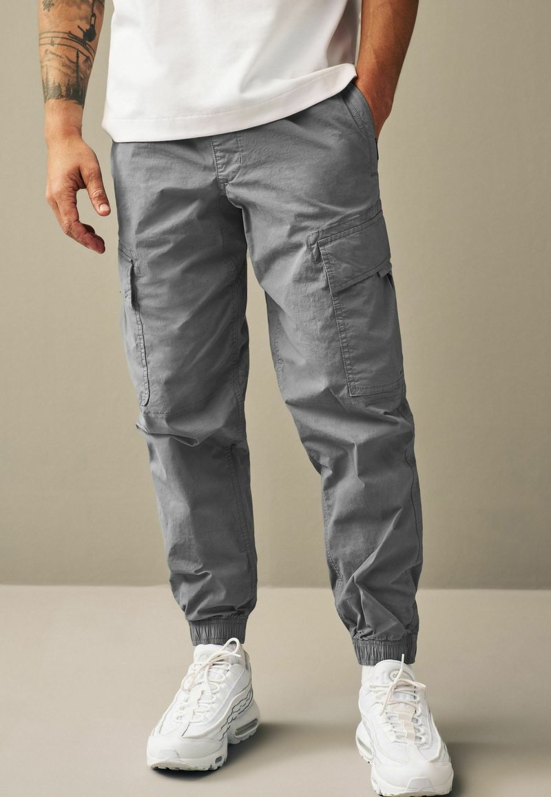 Pantaloni de marfă gri cu manșete elastice și buzunare laterale, fabricați dintr-o țesătură ușoară. Asortați cu adidași albi.