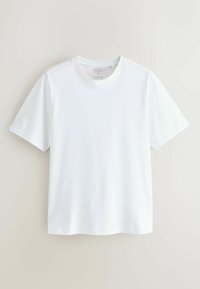 Lihtne valge lühikeste varrukatega T-särk, keskmine suurus, riputatud lamedalt helebeeži tausta vastu.