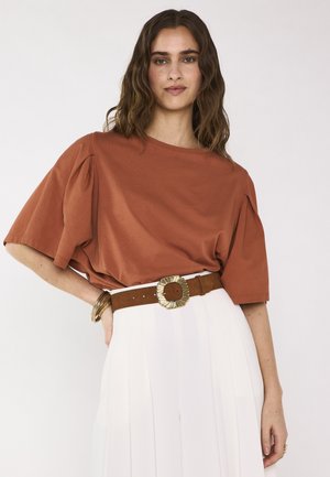 Femme aux cheveux bruns ondulés portant un haut ample couleur rouille, une jupe plissée blanche, une ceinture marron avec boucle dorée et des bracelets dorés.