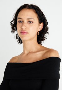 Fekete, vállnál nyitott top, texturált kötött mintával. A modellen göndör haj van, és kis arany fülbevalókat visel, fehér háttér előtt.