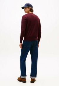 Suéter burdeos de cuello redondo, jeans rectos azul oscuro, zapatos marrones con puños enrollados y una gorra navy. Vista trasera con un fondo claro.