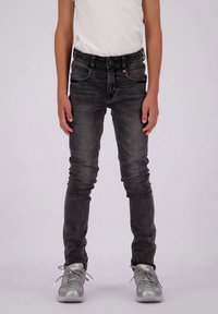 Dijete u iznošenim crnim skinny trapericama, bijeloj majici i sivim tenisicama, stoji uz jednostavnu svijetlu pozadinu.