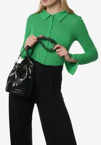 Borsa a secchiello nera in pelle stampata coccodrillo con doppi manici, hardware metallico e chiusura a coulisse, abbinata a una camicetta verde con colletto e pantaloni neri.