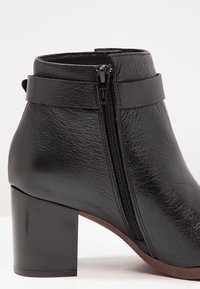 Bottine en cuir noir avec fermeture éclair sur le côté, talon bloc et détail de sangle près du haut, présentée sur un fond uni.