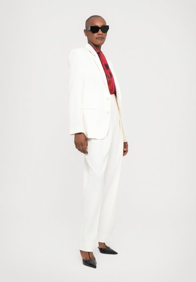 Witte op maat gemaakte pak met een blazer met twee knopen en brede broekspijpen, gecombineerd met een rode gestreepte stropdas en zwarte zonnebril. Puntige zwarte hakken.
