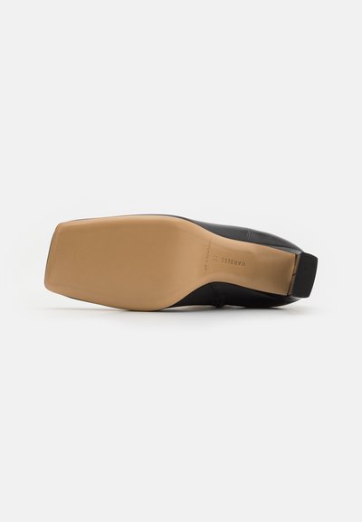 Black leather shoe with a rectangular toe, flat tan sole, and smooth texture. Markings on the sole indicate brand and country of manufacture.