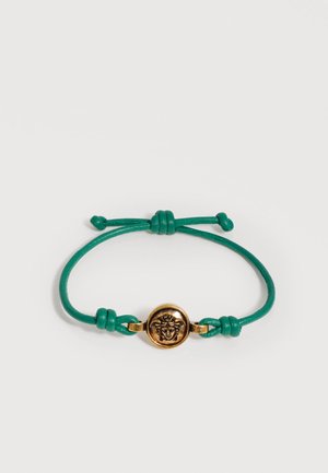 Bracelet à cordon vert ajustable avec un emblème tête de Méduse doré au centre, sur fond blanc uni.