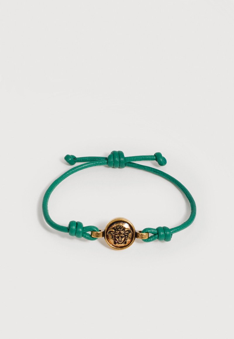 Bracelet à cordon vert ajustable avec un emblème tête de Méduse doré au centre, sur fond blanc uni.