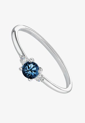 Anillo de plata con una piedra central redonda de color azul profundo, flanqueada por dos piedras claras más pequeñas, con una banda pulida y elegante.