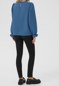 Blaue Bluse mit langen, gepufften Ärmeln, Schlüsselloch-Rückenansicht und strukturiertem Stoff, kombiniert mit schwarzen Slim-Fit-Jeans und schwarzen Plateauschuhen.