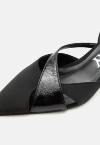 Γυναικείο παπούτσι με μαύρη μυτερή μύτη και λουριά από δέρμα λάκας και ύφασμα σε διάταξη σταυρού, σε λευκό φόντο.