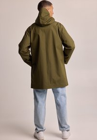 Olive green waterproof parka with a hood, featuring a loose fit, long sleeves, and a straight hem, paired with light blue jeans and white sneakers.