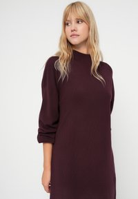 Abito in maglia bordeaux con scollatura alta e maniche a tre quarti. Texture liscia, vestibilità ampia, che arriva a metà coscia, design minimalista.