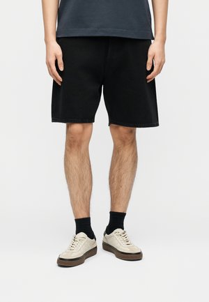 Mann trägt schwarze knielange Shorts, schwarze Socken und beige Turnschuhe, steht vor einem einfarbigen hellen Hintergrund.