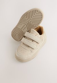 Beigetärryiset lenkkitossut, joissa on kaksi tarranauhaa, rei'itetty kärki ja kuvioitu kumipohja. Pehmeä vuori ja teksturoidut yläosan yksityiskohdat.