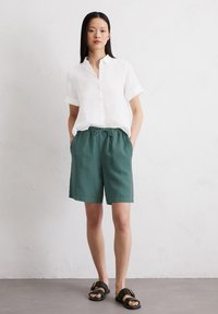 Chemise blanche à manches courtes avec boutons, associée à un short en lin vert et des sandales noires avec une boucle dorée. Coupe simple et décontractée.