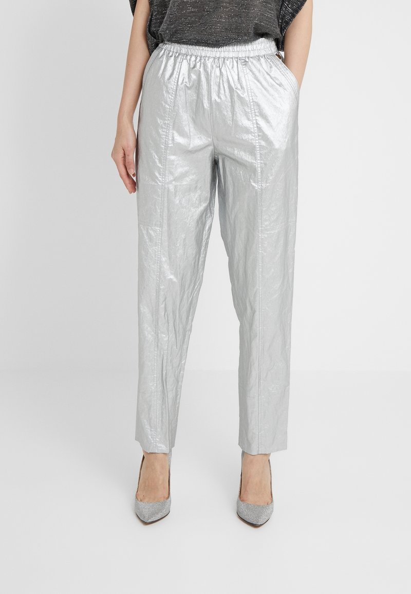 Personne portant un pantalon argenté brillant taille haute avec une ceinture élastique et des talons pointus argentés assortis, debout les mains dans les poches.