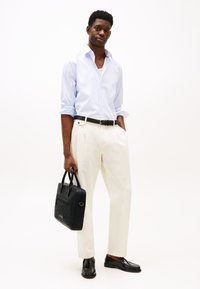 Light blue striped shirt, cream trousers, black belt and shoes, holding a black leather bag with a textured pattern.