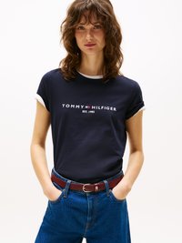Femme aux cheveux bruns bouclés portant un t-shirt navy de Tommy Hilfiger, un jean bleu et une ceinture rouge foncé, se tenant les mains dans les poches.