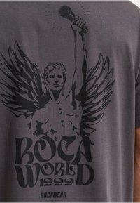 Pelēka kokvilnas T-kreklu ar melnu grafiku, kurā attēlota plika figūra ar spārniem, kas tur mikrofonu; apakšā ir teksts "ROCA WORLD 1999".