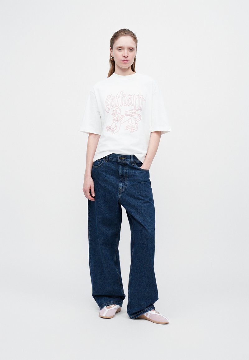 Ungdommelig kvinne iført en hvit oversized T-skjorte med et lion-motiv, løse mørkeblå jeans og lyse rosa sko med hvite reimer.
