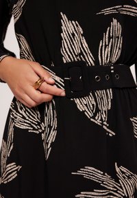 Czarna sukienka z liściastym, beżowym wzorem, z szerokim czarnym paskiem z tkaniny i prostokątną klamrą oraz oczkami. Ręka oparta na pasku.