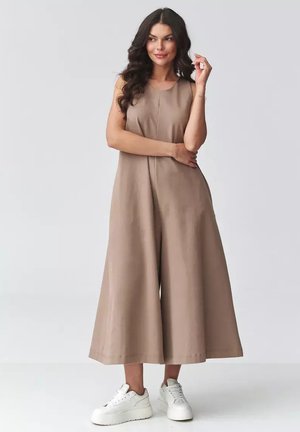 Kobieta o długich ciemnych włosach, ubrana w bezrękawnikowy, beżowy kombinezon z szerokimi nogawkami oraz białe sneakersy na platformie, stojąca na jednolitym tle.