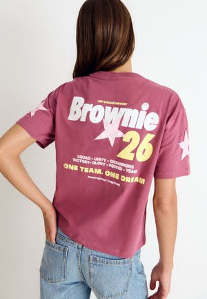 Persona con cabello largo y castaño, que lleva una camiseta burdeos con estrellas y texto "Brownie 26" y vaqueros azul claro, vista desde atrás.