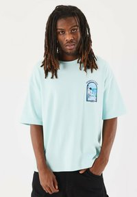 Camiseta de gran tamaño de color azul claro con un estampado gráfico en el lado izquierdo del pecho. Mangas cortas, textura suave, hecha de una mezcla de algodón.