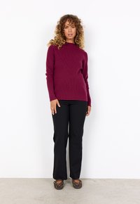 Burgunderfarbener, gerippter Strickpullover, schwarze, enganliegende Hosen und braune Schuhe mit goldenen Akzenten. Das Outfit zeigt eine einfache, klar strukturierte Silhouette.