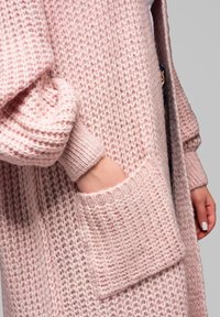 Růžový pletený kardigan s texturovaným vzorem, který má velkou kapsu a manžety s pružným lemem. Měkká, nadýchaná tkanina poskytuje teplo a pohodlí.