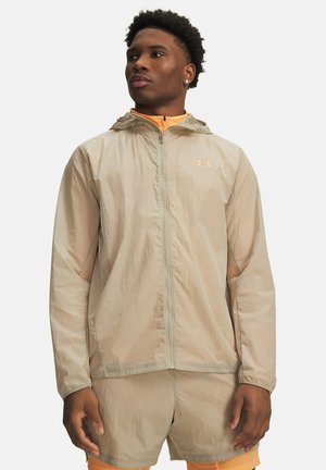 Lightweight beige windbreaker jacket with hood, featuring a front zipper, elastic cuffs, and subtle logo. Paired with orange athletic shorts.