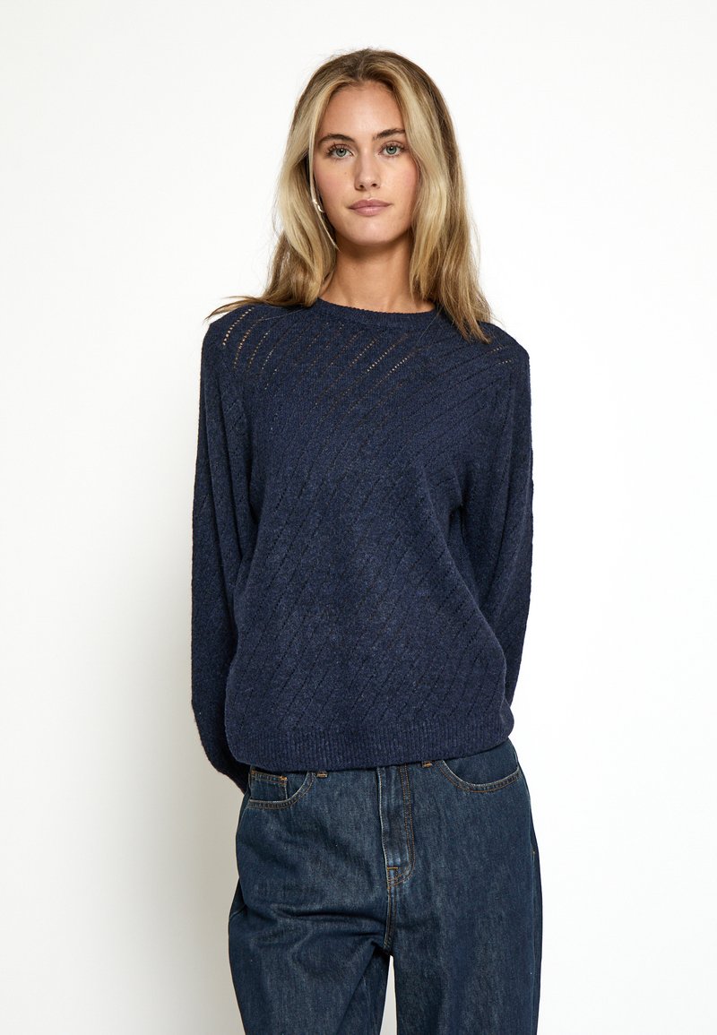 Marineblå strikket sweater med et tekstureret diamantmønster, rund halsudskæring og lange ærmer, parret med løstsiddende mørke denimjeans.