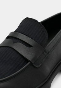 Μαύρο δερμάτινο μοκασίνι με επάνω μέρος από υφή ναυτικού μπλε υφάσματος, που διαθέτει καμπυλωτή μαύρη λωρίδα ως διακοσμητικό στο μπροστινό μέρος. Ομαλή υφή και στρογγυλεμένο σχήμα.