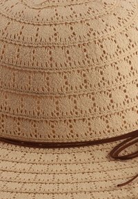 Beżowy pleciony kapelusz z warstwowym wzorem, z okrągłymi perforacjami i brązową wstążką wokół podstawy. Lekki i teksturowany materiał.