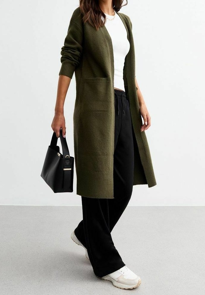 Cardigan lungo verde oliva con polsini a coste e due tasche anteriori, abbinato a pantaloni neri e sneaker bianche, mentre tiene una borsa nera.