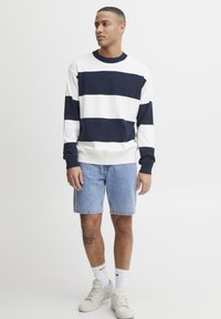 Maglione a righe blu navy e bianche con scollo rotondo, abbinato a pantaloni corti di denim. Sneakers e calze completano l'outfit casual.