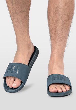 Pies con sandalias tipo slip-on de color gris oscuro con suela texturizada y logo "HH" en las correas sobre un fondo liso y claro.