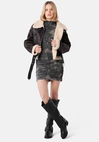 Brown leather jacket with a cream shearling collar, worn over a fitted, textured, dark camouflage dress. Knee-high black leather boots with straps.